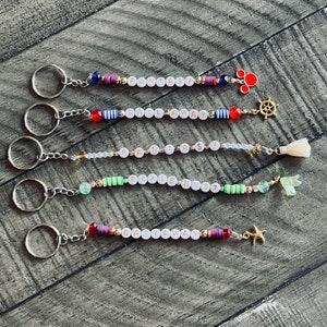 May include: Five colorful beaded keychains with different charms. The keychains spell out "Fantasy", "Sail Away", "Fantasy", "Pixie Dust", and "Castaway".