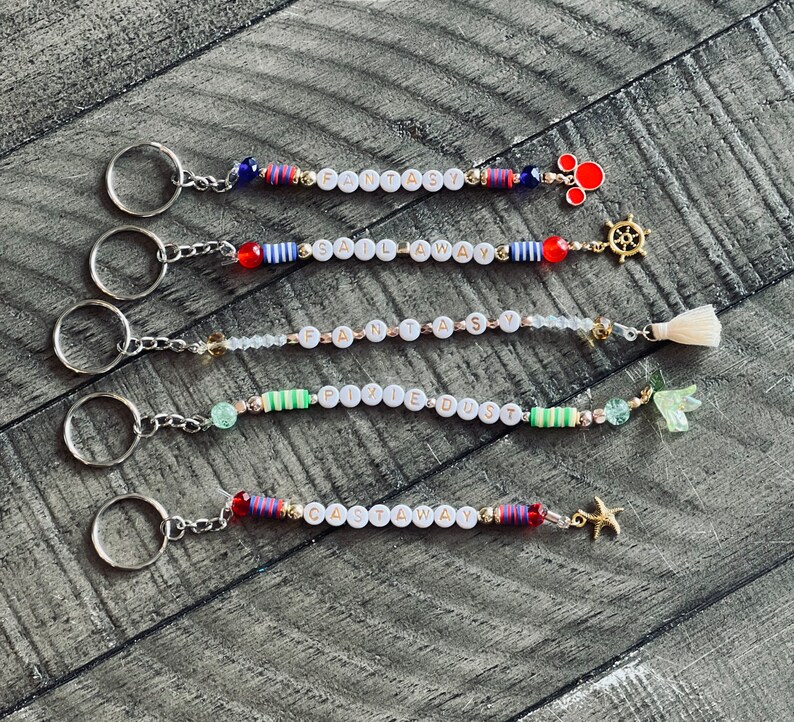 May include: Five colorful beaded keychains with different charms. The keychains spell out "Fantasy", "Sail Away", "Fantasy", "Pixie Dust", and "Castaway".