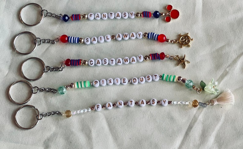May include: Five keychains with different colored beads and charms. The keychains spell out the words "Fantasy", "Sail Away", "Castaway", "Pixie Dust", and "Fantasy".