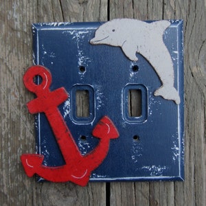 May include: A blue and white light switch cover with a red anchor and a white dolphin. The anchor is on the left side of the cover and the dolphin is on the right side. The cover has a distressed look.