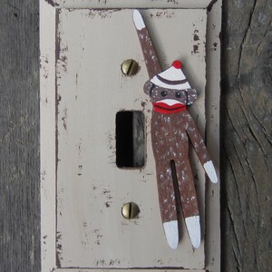 May include: A decorative light switch cover featuring a painted sock monkey design. The monkey is brown with white accents, a red mouth, and a white and striped hat. The cover is off-white with a distressed finish.