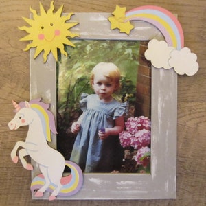 May include: A gray wooden picture frame with a whitewashed finish. The frame is decorated with a yellow sun, a rainbow, a cloud, and a unicorn. A photo of a young girl in a blue dress is in the frame.