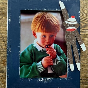 May include: A blue picture frame with a white distressed finish. The frame holds a photo of a young child wearing a green sweater and eating a lollipop. A wooden sock monkey is attached to the frame.