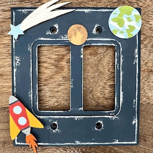 May include: A black double light switch cover with a white distressed finish. The cover is decorated with a yellow and red rocket, a white comet, a blue and green planet, and an orange moon.