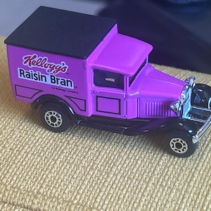 Może przedstawiać: Fioletowa ciężarówka zabawka z czarnym dachem i detalami. Ciężarówka jest oznaczona logo "Kellogg's Raisin Bran". Pojazd ma czarne koła i szczegółowy przedni grill.