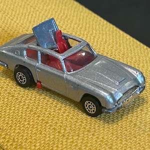 Puede incluir: Un coche de juguete plateado con interior rojo y techo azul que se abre. El coche tiene ruedas negras y está sobre una superficie amarilla. El coche es una réplica en miniatura de un coche deportivo clásico.