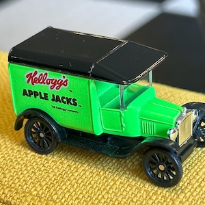 Puede incluir: Un camión de juguete verde y negro con el logotipo de Kellogg's Apple Jacks en el lateral. El vehículo de estilo vintage tiene ruedas negras y un techo negro. El camión de juguete está sobre una superficie amarilla.