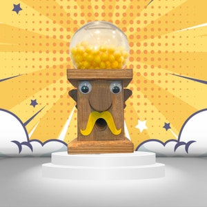 May include: A wooden gumball machine with a cartoon face, googly eyes, and a yellow mustache. The machine is filled with yellow gumballs and is sitting on a white platform.