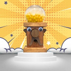 May include: A wooden gumball machine with a cartoon face, googly eyes, and a mustache. The machine is filled with yellow gumballs and is sitting on a white platform.