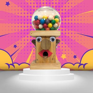 May include: A wooden gumball machine with a face, featuring googly eyes, a brown nose, and a red mouth. The gumball machine is filled with colorful gumballs and is sitting on a white pedestal.