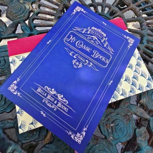 May include: A blue reading journal titled My Classic Books by Bella Boo & Books with white decorative borders and text. Beneath it lies a second book featuring a red spine and a cream-colored cover with a repeating blue fan-shaped pattern.