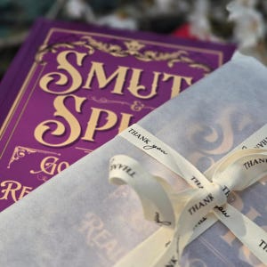 May include: A purple book with gold lettering reading "SMUT & SPY" is partially visible, wrapped in white paper and tied with a cream ribbon that says "Thank you". The book's cover has ornate gold designs.