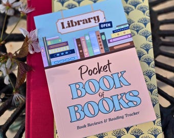 May include: A rectangular reading tracker booklet titled Pocket Book of Books with a light pink cover and a blue illustration of a library shelf. The text Library Open is visible at the top. It rests on a yellow patterned book and a red book.