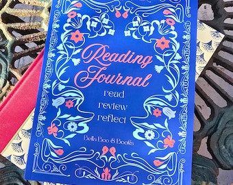 May include: A blue reading journal with the words "Reading Journal" in a cursive font. The journal has floral and vine designs in white, light blue, and coral. The text "read, review, reflect" is below the title, and the brand name "Bella Boo & Books" is at the bottom.