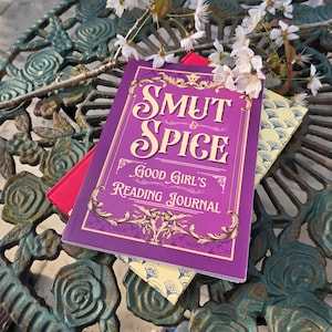 May include: A purple "Smut & Spice Good Girl's Reading Journal" with gold accents and text. A red book and a patterned yellow book are beneath it. White flowers and green metal table are in the background.