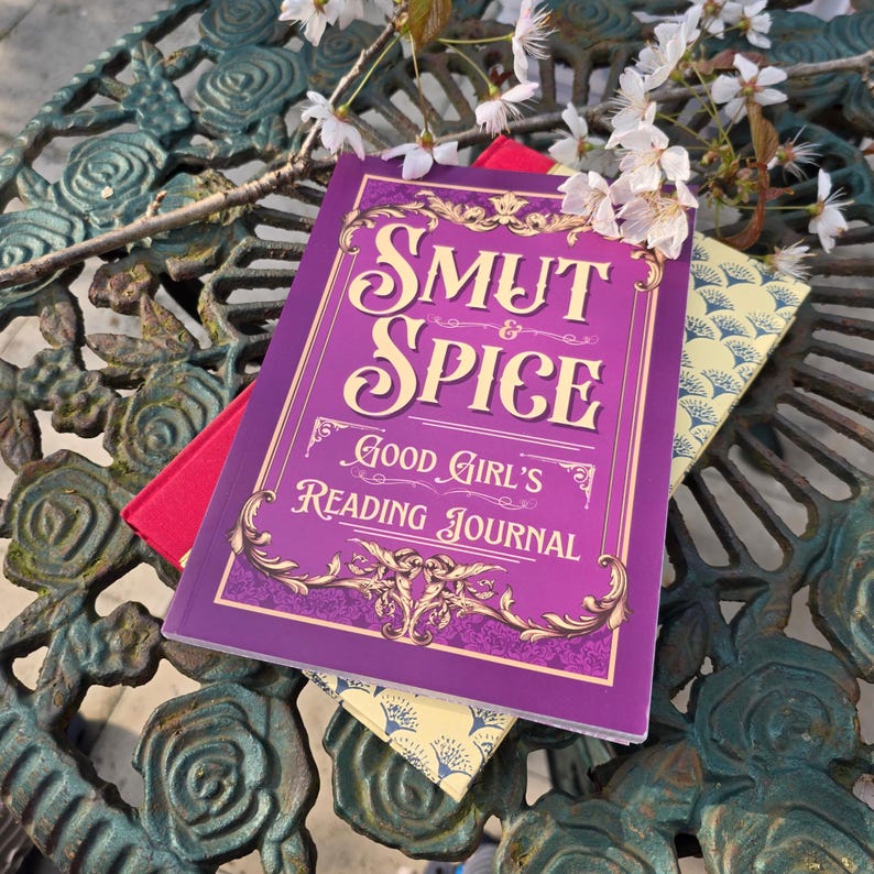 May include: A purple "Smut & Spice Good Girl's Reading Journal" with gold accents and text. A red book and a patterned yellow book are beneath it. White flowers and green metal table are in the background.