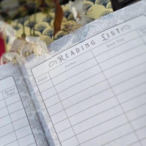May include: Open notebook with a "Reading List" page, featuring lines for author, book title, and dates. The notebook has a decorative floral pattern in gray and white. The text "Reading List" is at the top of the page.