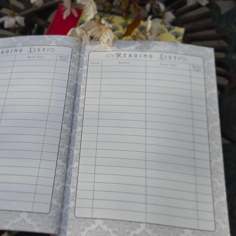 May include: Open notebook with lined pages for recording book titles, author, and review details. The notebook has a decorative border and the words "Reading List" at the top of each page. The background includes white flowers and a red book.