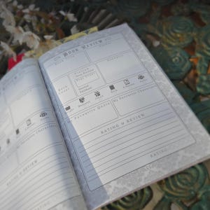 May include: Open book review journal with a floral patterned border. The journal has sections for book title, author, synopsis, and rating. Includes icons for audio, e-reader, and book.