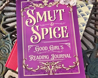 May include: A purple journal with the words "SMUT & SPICE" and "GOOD GIRL'S READING JOURNAL" in white script. The journal has decorative gold accents and is surrounded by other books and white flowers.