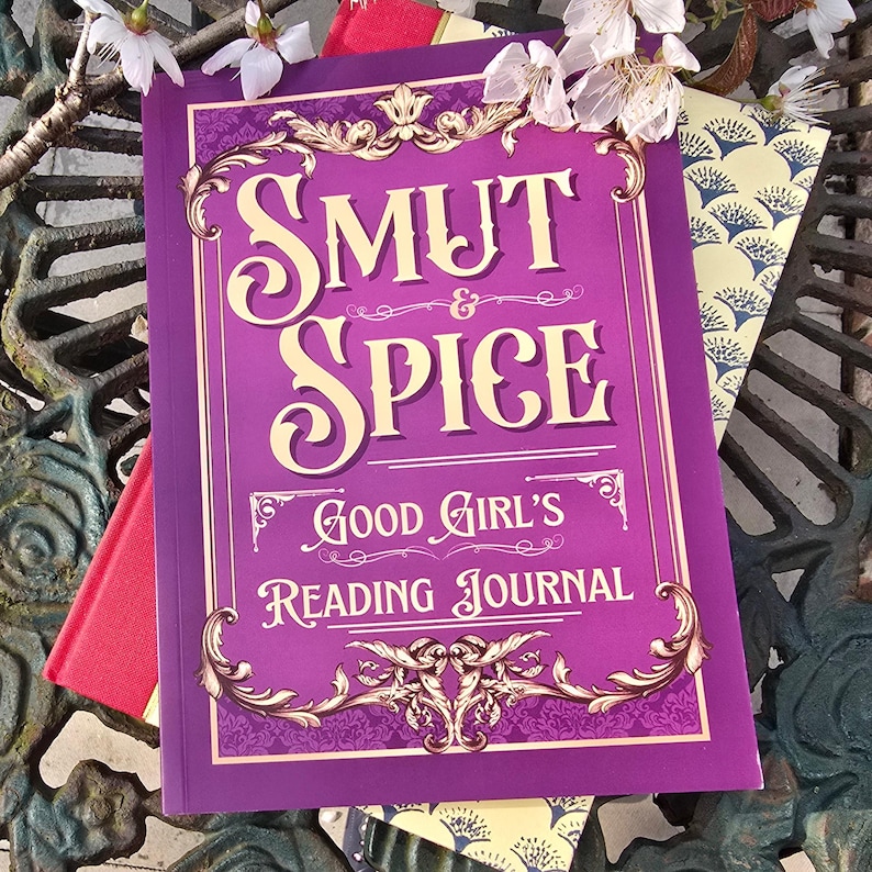 May include: A purple journal with the words "SMUT & SPICE" and "GOOD GIRL'S READING JOURNAL" in white script. The journal has decorative gold accents and is surrounded by other books and white flowers.