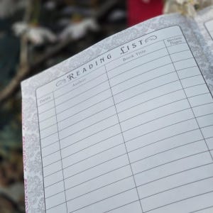 May include: Open notebook with a "Reading List" title. The pages are white with gray lines and a decorative border. The notebook is open to a page with spaces for book titles, authors, dates, and review pages.