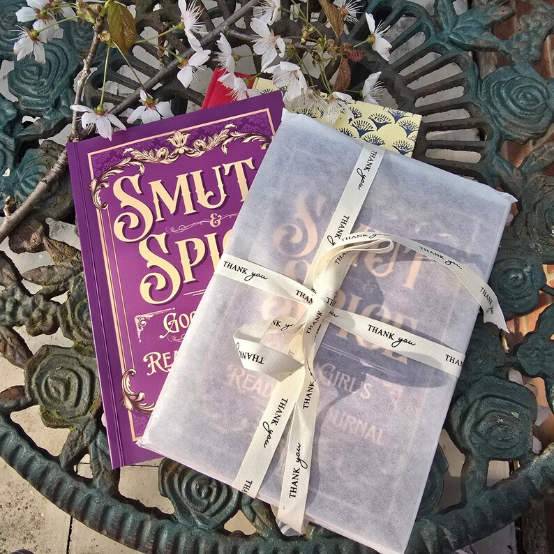 May include: Two books, one wrapped in white paper and tied with a ribbon that says "Thank you." The other book is purple with gold lettering that reads "Smut & Spice." The books are on a decorative metal surface.