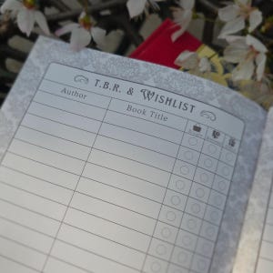 May include: Open notebook page with the title "T.B.R. & Wishlist" and sections for author and book title. The page has a decorative floral pattern and checkboxes for tracking book status. A red book is visible in the background.