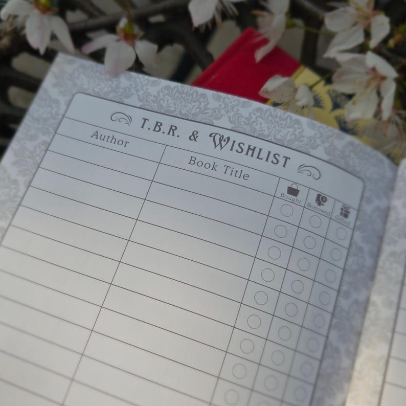 May include: Open notebook page with the title "T.B.R. & Wishlist" and sections for author and book title. The page has a decorative floral pattern and checkboxes for tracking book status. A red book is visible in the background.