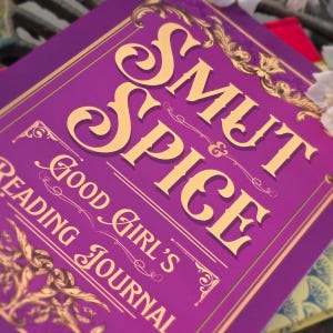 May include: A purple journal with gold lettering that reads "Smut & Spice Good Girl's Reading Journal." The cover has ornate gold decorative elements. The journal is designed for reading and journaling.