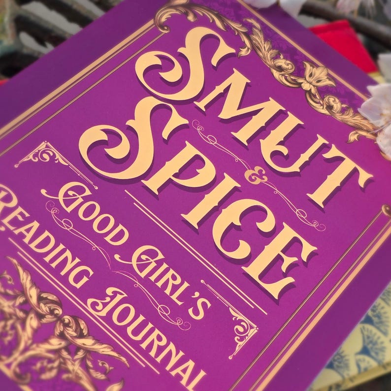 May include: A purple journal with gold lettering that reads "Smut & Spice Good Girl's Reading Journal." The cover has ornate gold decorative elements. The journal is designed for reading and journaling.