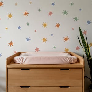 Op de afbeelding: Een commode van licht hout met een zachtroze aankleedkussen staat tegen een muur die is versierd met verspreide stervormige muurstickers in gedempte tinten oranje, geel, blauw, groen en roze, wat zorgt voor een kleurrijk en patroonrijk geheel.