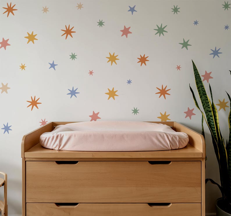 Op de afbeelding: Een commode van licht hout met een zachtroze aankleedkussen staat tegen een muur die is versierd met verspreide stervormige muurstickers in gedempte tinten oranje, geel, blauw, groen en roze, wat zorgt voor een kleurrijk en patroonrijk geheel.