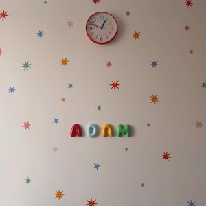 Op de afbeelding: Een wanddecoratie met een ronde rode analoge klok met zwarte cijfers en blauwe wijzers, geplaatst boven de naam ADAM, gespeld in kleurrijke, driedimensionale, afgeronde letters. De muur is versierd met verspreide, veelkleurige sterstickers.