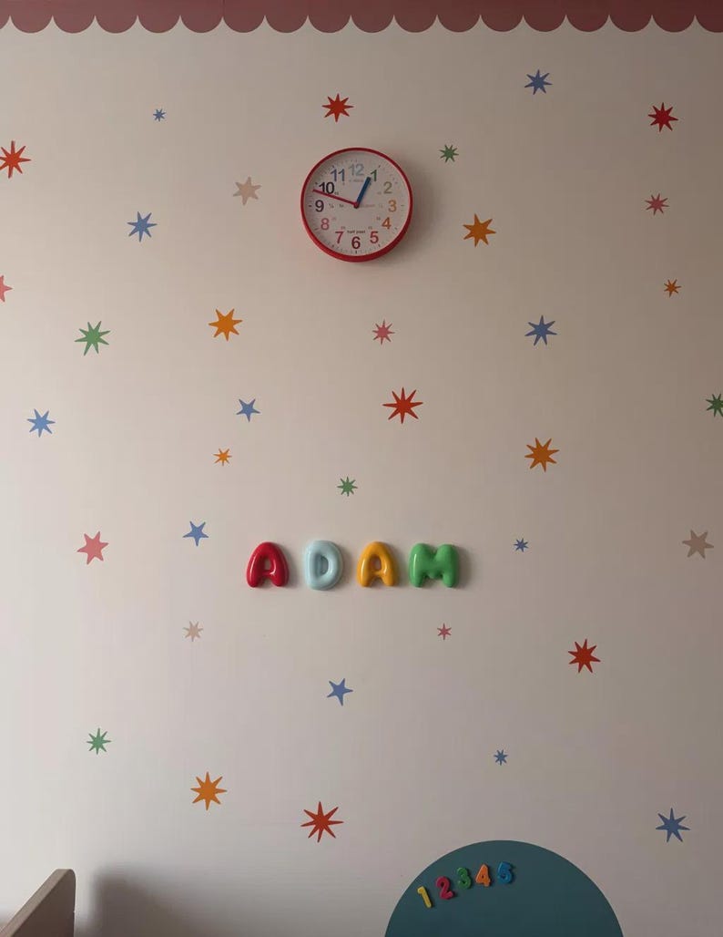 Op de afbeelding: Een wanddecoratie met een ronde rode analoge klok met zwarte cijfers en blauwe wijzers, geplaatst boven de naam ADAM, gespeld in kleurrijke, driedimensionale, afgeronde letters. De muur is versierd met verspreide, veelkleurige sterstickers.