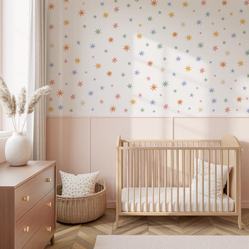 Op de afbeelding: Een muur bedekt met wit behang met een herhalend patroon van kleine, veelkleurige sterretjes in tinten oranje, blauw, groen en roze, geplaatst boven een lichtroze lambrisering in een kamer met een houten babybedje.