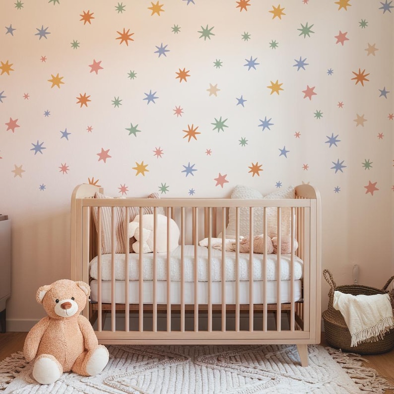 Op de afbeelding: Een lichthouten ledikant met een wit matras, een beige teddybeer en een wit, getextureerd kleed. De muur achter het ledikant is voorzien van een patroon van kleine, veelkleurige sterstickers in oranje, blauw, groen, geel en roze.