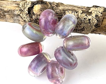 May include: A set of eight glass beads in shades of pink, purple, and white with iridescent swirls. The beads are donut shaped and have a glossy finish.