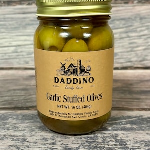May include: A glass jar filled with green garlic stuffed olives. The jar has a brown label with the text "DADDINO Family Farm" and "Garlic Stuffed Olives". The label also includes the net weight of 16 ounces (454g).
