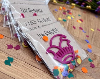 May include: Several packages with the text "TEN DONDRIA" and a pink floral-shaped object are visible. The packages are sealed in clear plastic and contain colorful confetti. The packages are on a wooden surface.