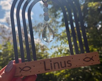 May include: A decorative wooden rainbow with the name "Linus" engraved on the base. The rainbow has black arches and a clear crystal teardrop pendant. The design includes a fish symbol.