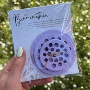 Könnte beinhalten: Eine Verpackung mit drei lavendelfarbenen, kreisförmigen Blumenarrangiergittern mit mehreren Löchern. Die Verpackung enthält eine Karte mit dem Text "Die Blumenretterin" und zusätzlichem deutschem Text. Die Gitter sind mit einem naturfarbenen Bindfaden zusammengebunden.