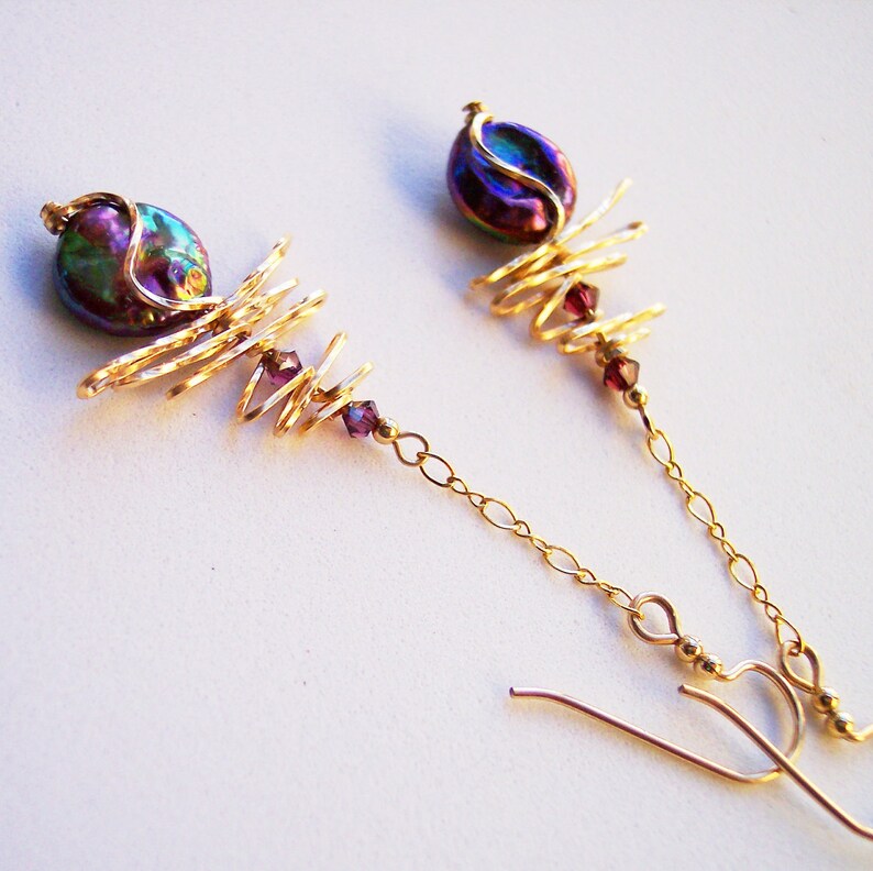 May include: A pair of gold-coloured earrings. Each earring has an iridescent, oval bead at the top, with a spiral wire design and small purple beads. The earrings hang from a delicate gold chain.