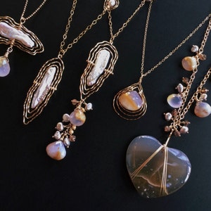 May include: Several gold necklaces with diverse pendant styles. Some pendants have large, irregular pearls, others feature teardrop-shaped stones and a heart-shaped stone. The necklaces are presented against a black backdrop.