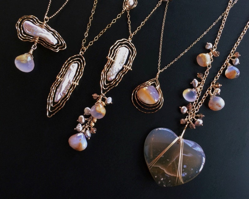 May include: Several gold necklaces with diverse pendant styles. Some pendants have large, irregular pearls, others feature teardrop-shaped stones and a heart-shaped stone. The necklaces are presented against a black backdrop.
