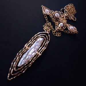May include: A gold-coloured necklace featuring a large, elongated pearl pendant with a layered, openwork design. The necklace also includes smaller pearl accents and delicate chain detailing, all set against a dark background.