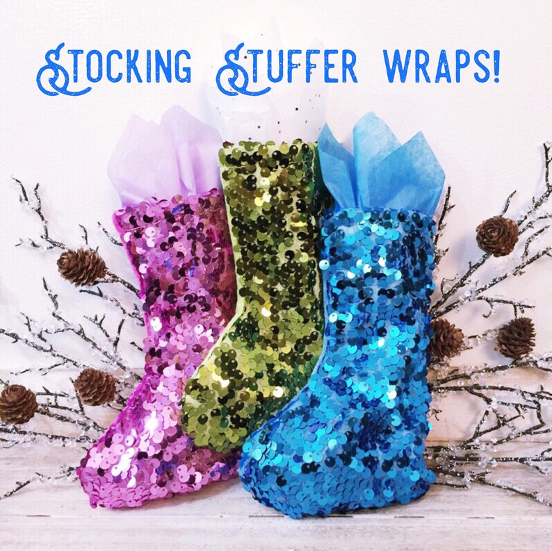May include: Three sequined stocking-shaped gift wraps in pink, green, and blue, each filled with tissue paper. The words "STOCKING STUFFER WRAPS!" are written in blue script above the stockings. Decorative branches with pinecones are on either side.