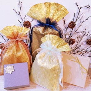 May include: A selection of gift bags and boxes in gold, tan, and lavender. The bags feature a textured, pleated design and are tied with ribbons in various colours. A small lavender box with a snowflake decoration is also present.