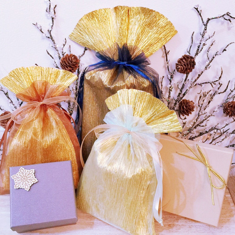 May include: A selection of gift bags and boxes in gold, tan, and lavender. The bags feature a textured, pleated design and are tied with ribbons in various colours. A small lavender box with a snowflake decoration is also present.