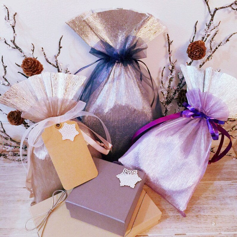 May include: A selection of gift bags and boxes in silver, lavender, and beige. The bags are made of shimmering fabric and tulle, tied with ribbons. Some bags have decorative tags with snowflake accents. Small pinecones and branches are in the background.